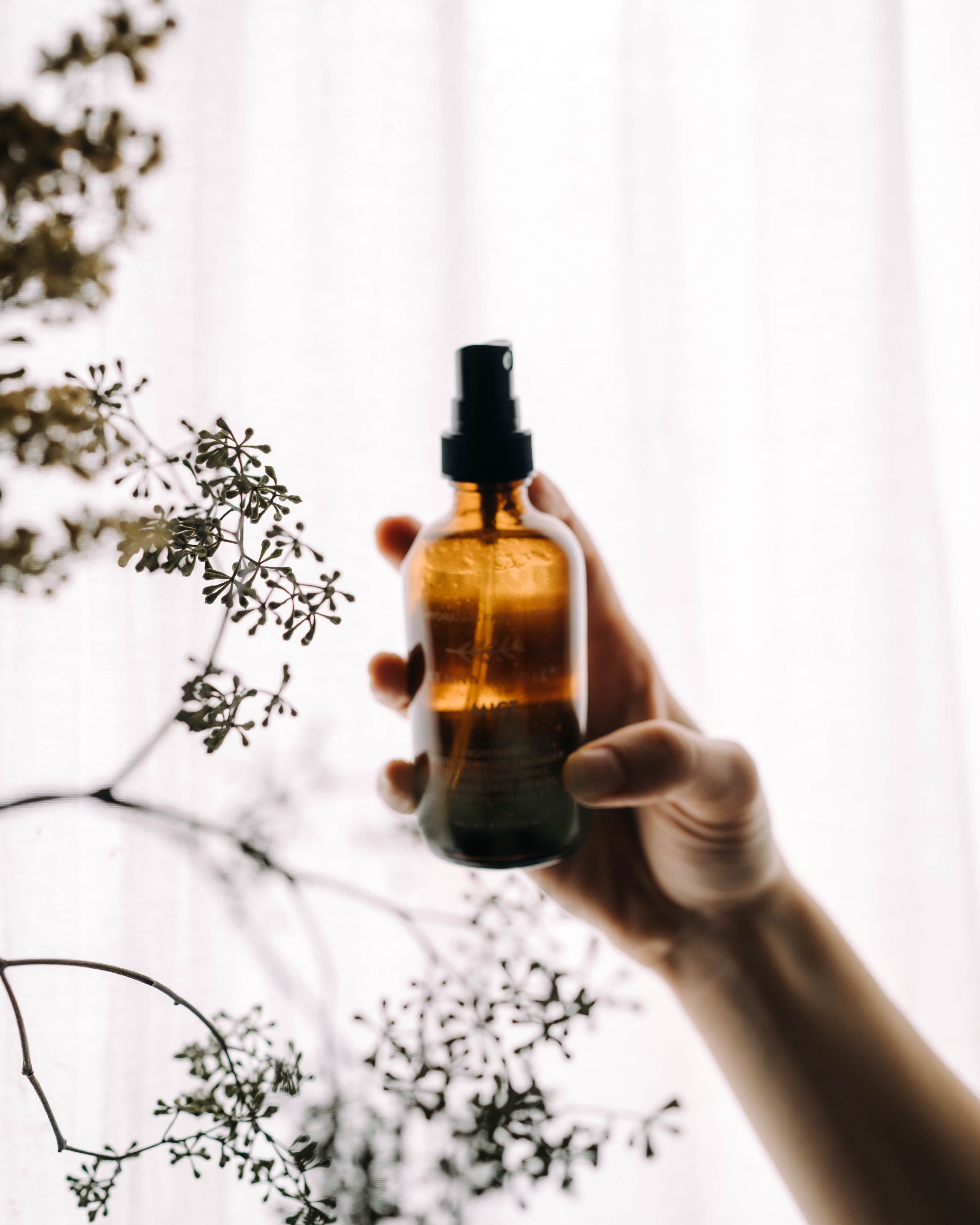 Hand holding an amber glass bottle with a black spray nozzle against a blurred natural background